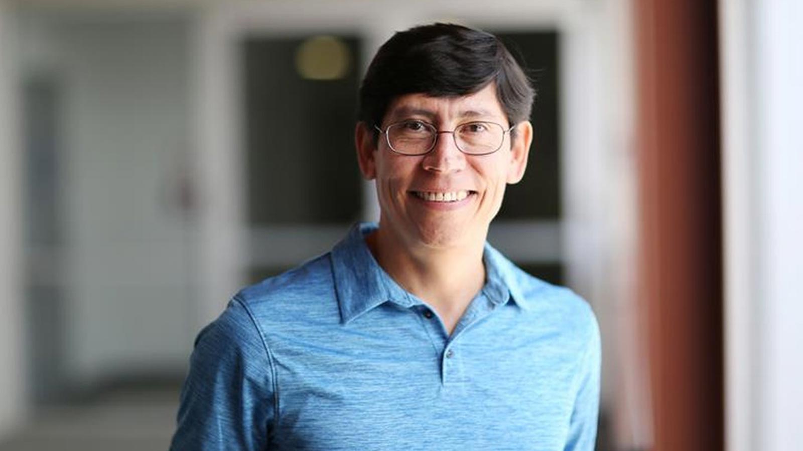 Cal State LA professor Arturo Pacheco-Vega smiles in a portrait taken in a campus hallway, wearing a blue collared shirt.