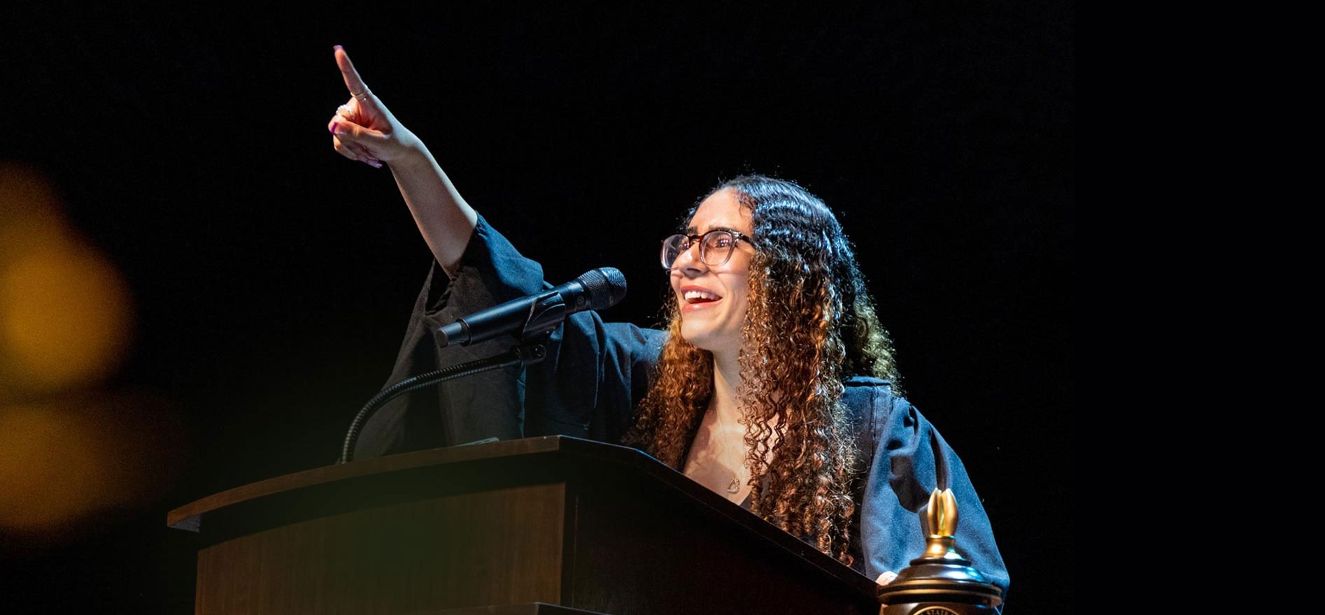Cal State LA graduate Arwa Hammad speaks at the podium during Commencement, smiling and pointing upward while addressing the audience.