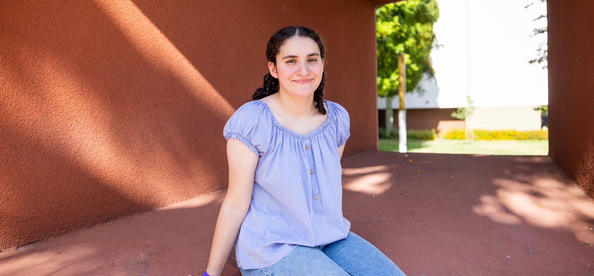 Cal State LA student Briana Zavala sits outside a campus building, smiling in natural sunlight with trees and architecture in the background.