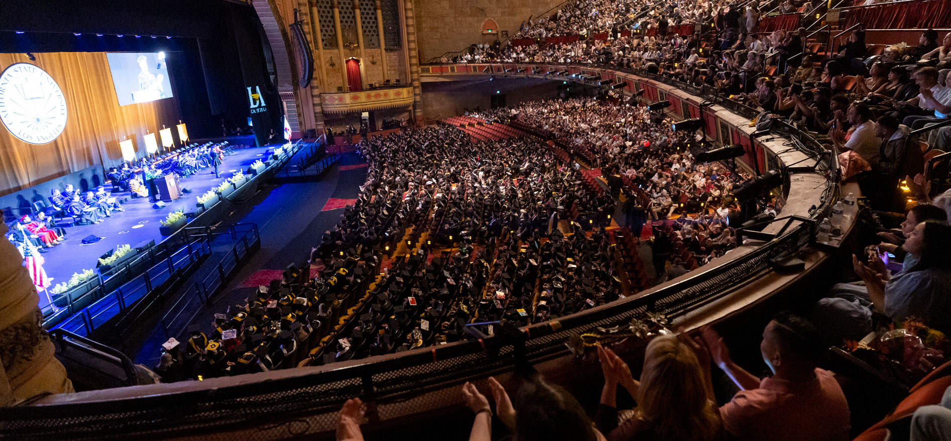 Cal State LA graduates and guests fill the Shrine Auditorium during the 2025 Commencement ceremony, with University leaders speaking on stage under the Cal State LA seal.