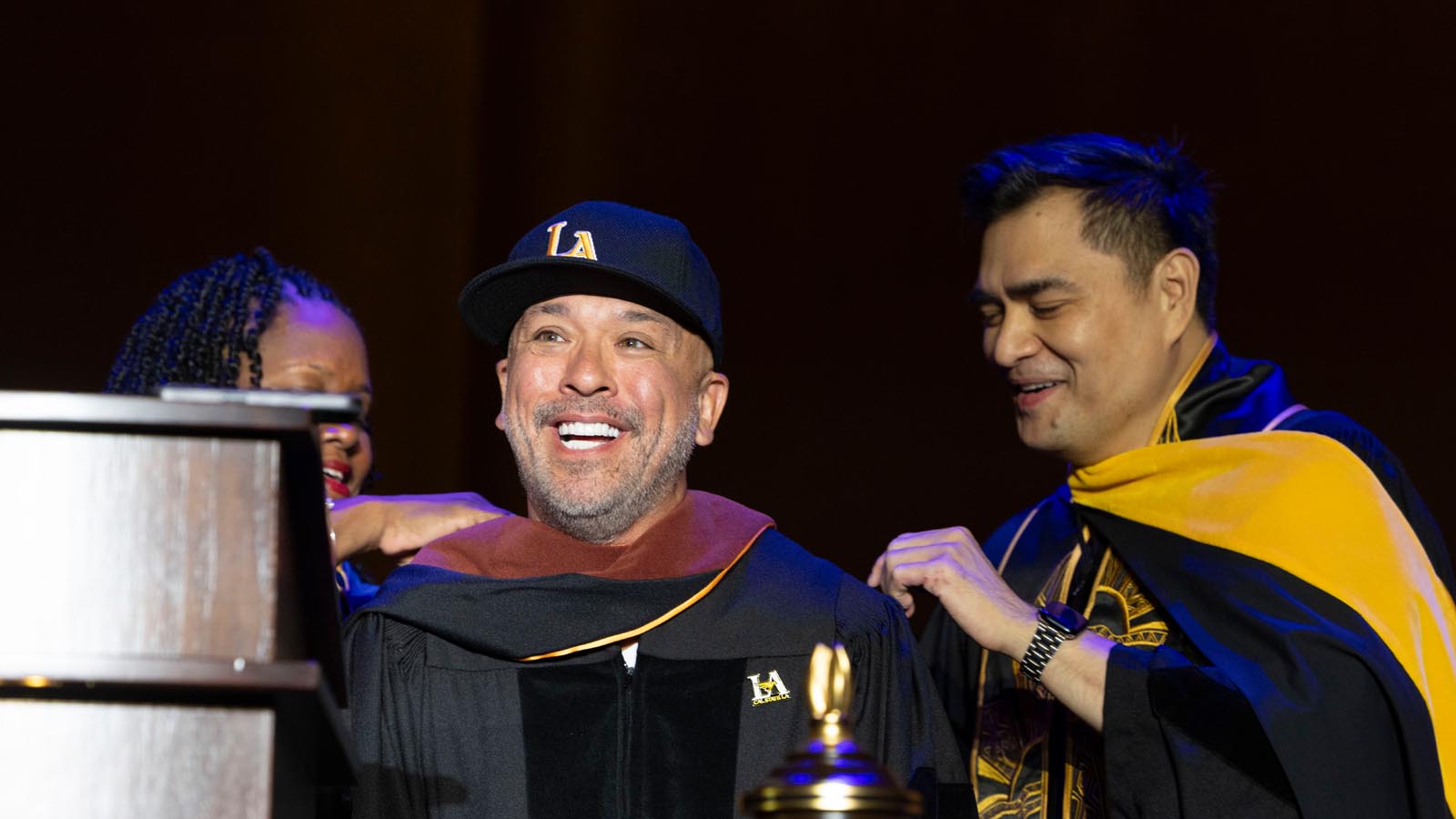 Cal State LA President Berenecea Johnson Eanes helps comedian Jo Koy don academic regalia after receiving an honorary degree during the 2025 Commencement ceremony.