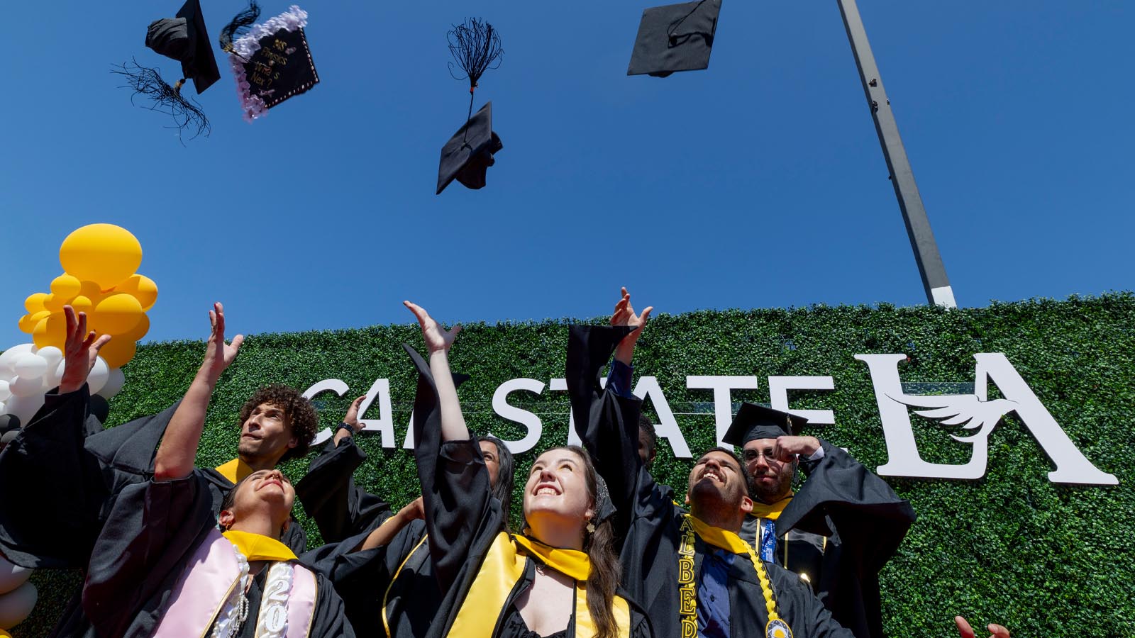 Cal State LA graduates toss their caps into the air in celebration during the May 2025 Commencement ceremony, standing in front of a Cal State LA backdrop with balloons.