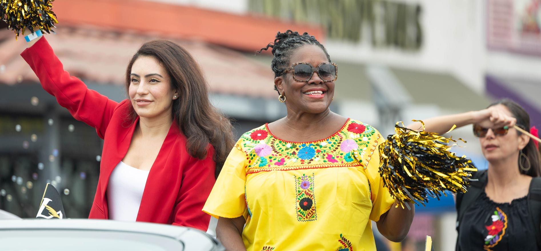 Cal State LA President Berenecea Johnson Eanes waves and smiles while riding in a parade, wearing a bright yellow embroidered top and holding black and gold pom-poms alongside community participants.
