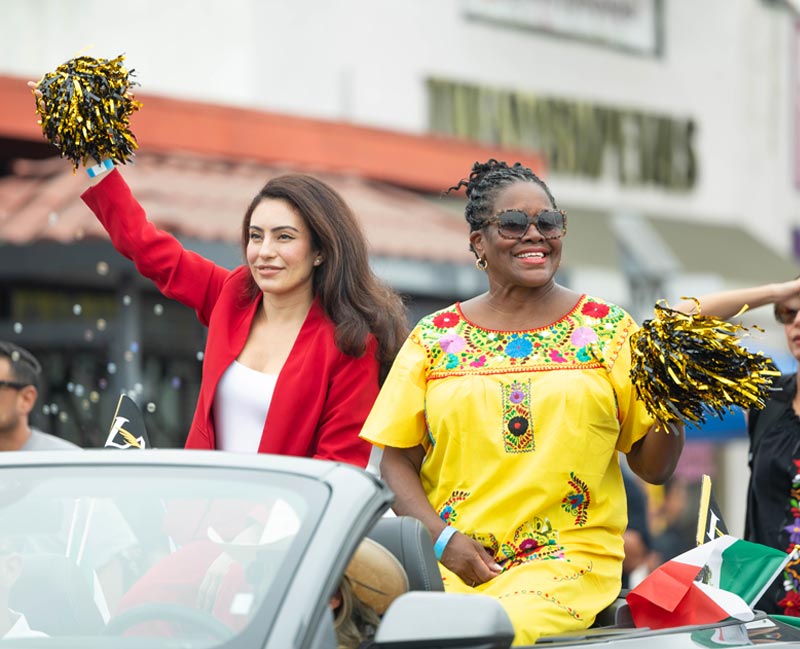 Two people riding in the back of a car during a parade.