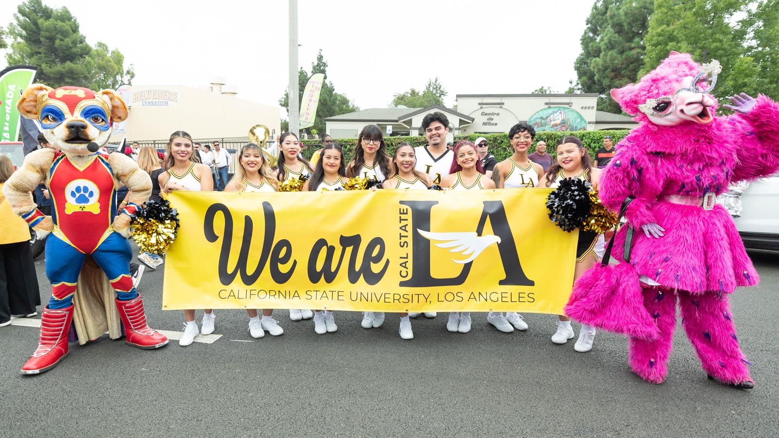 Cal State LA cheer team marches in a community parade holding a bright yellow “We are LA” banner alongside university mascots.
