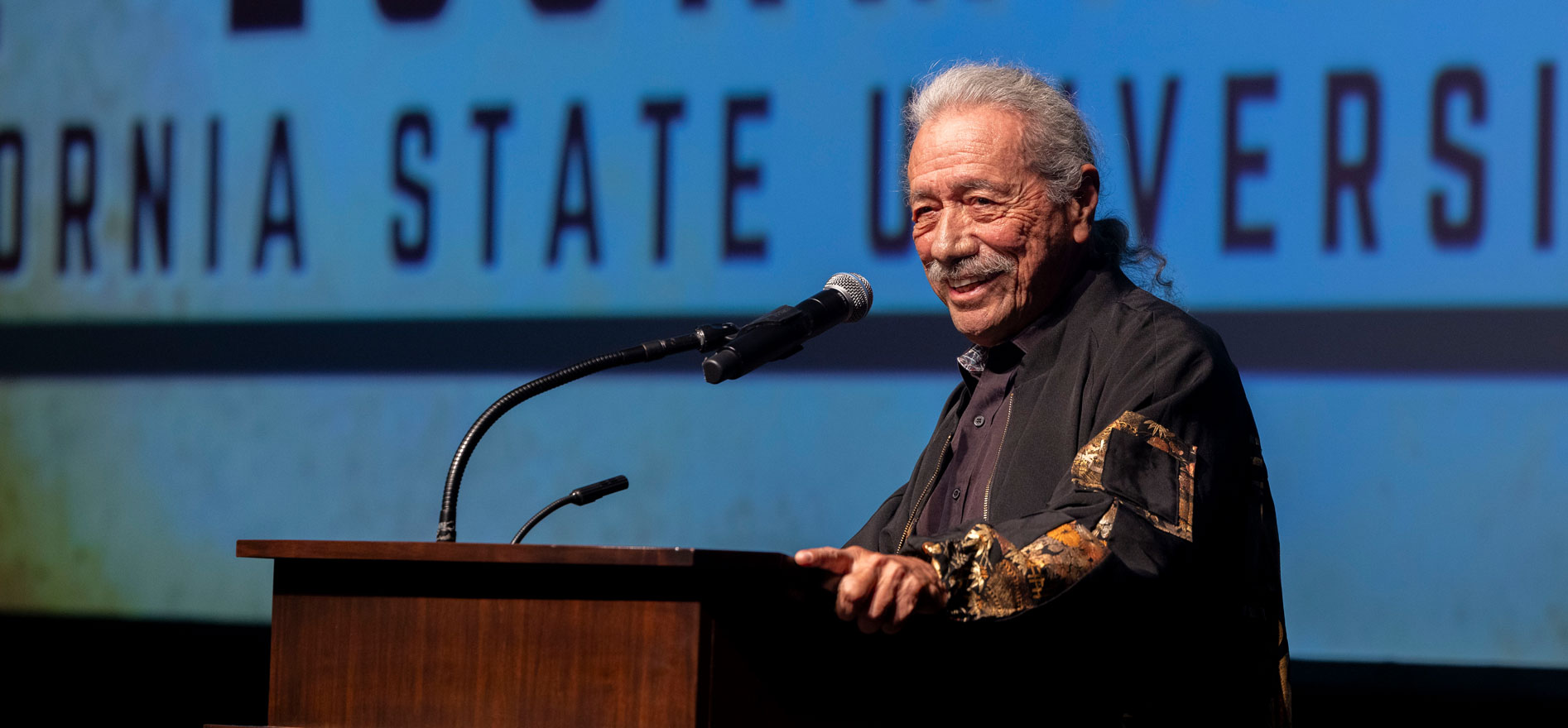 Actor and activist Edward James Olmos speaks at the Cal State LA podium during a film screening event, smiling as he addresses the audience.