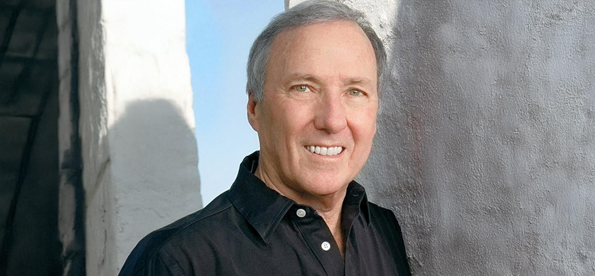 Portrait of Cal State LA alumnus and author Joseph Wambaugh, wearing a black shirt and smiling while standing against a light-colored wall.