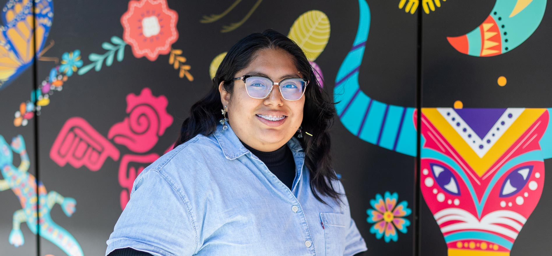 Cal State LA student Maria Velasco stands in front of a colorful mural featuring bright abstract animal and floral designs on campus