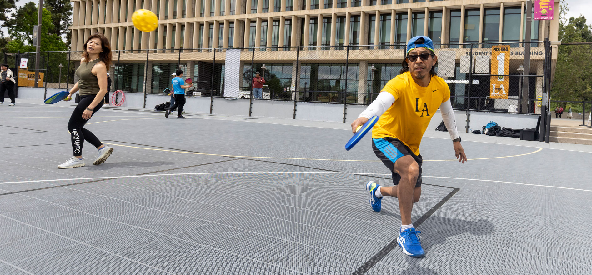 Two people play pickleball on an outdoor court at Cal State LA, with one player in a yellow Cal State LA shirt lunging to hit the ball near the Student Services Building.