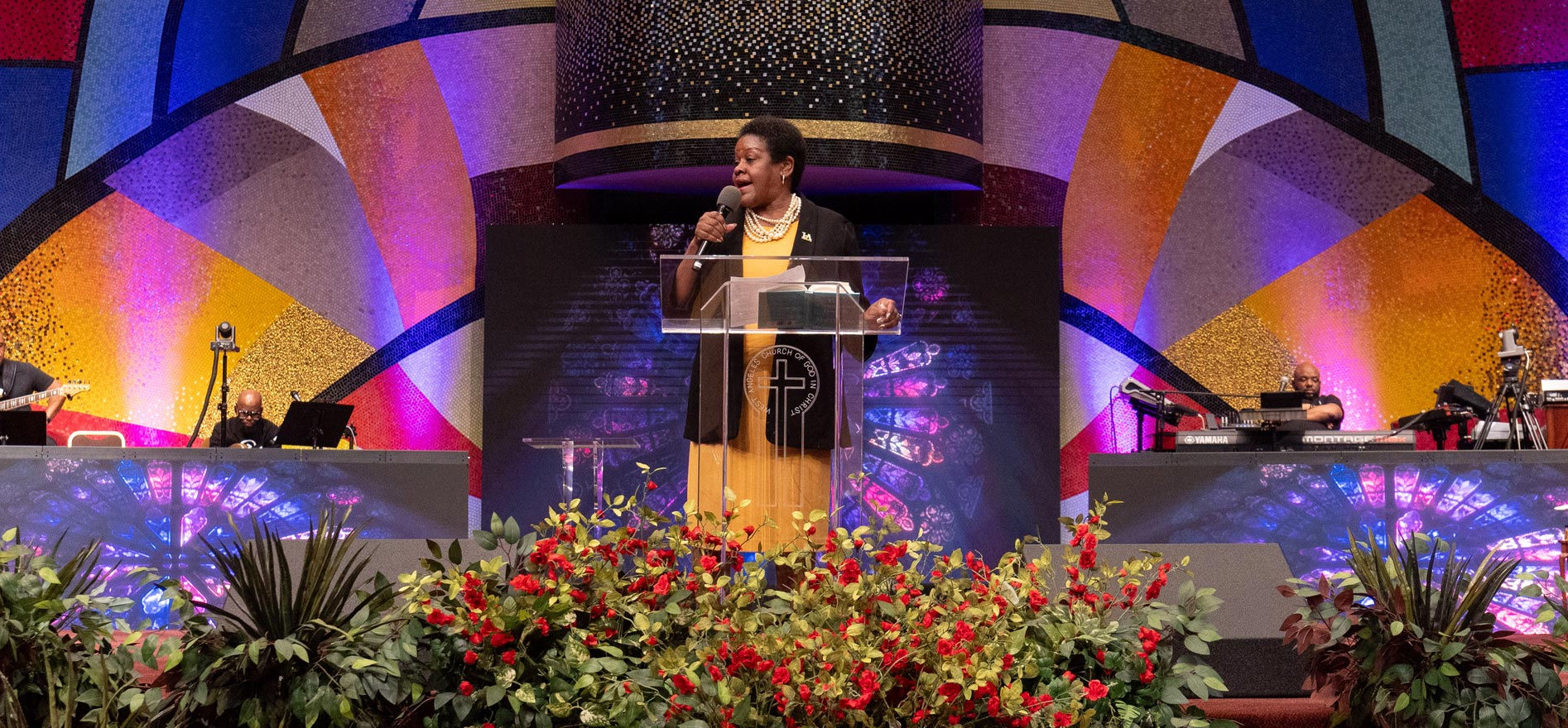 Cal State LA President Berenecea Johnson Eanes speaks at the podium during CSU Super Sunday, addressing a congregation in a church adorned with colorful stained-glass designs and floral arrangements.