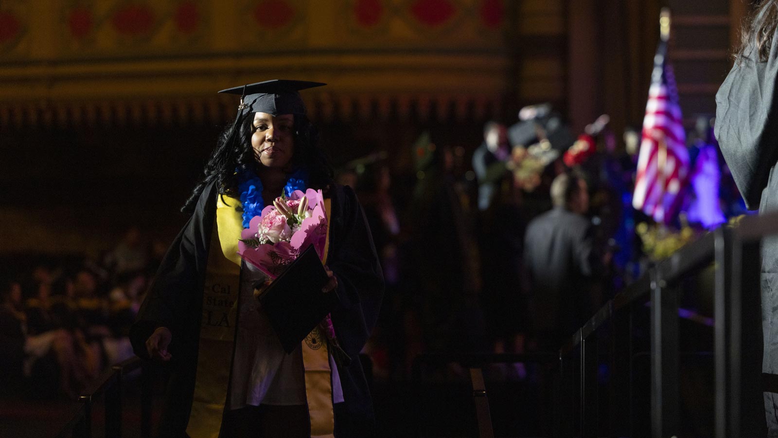 A Cal State LA graduate walks across the stage holding flowers and a diploma cover during the 2025 Commencement ceremony at the Shrine Auditorium.