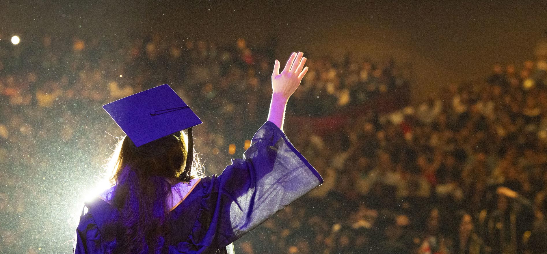 A Cal State LA graduate waves toward the audience from the stage during the 2025 Commencement ceremony, illuminated by stage lights and surrounded by cheering guests.