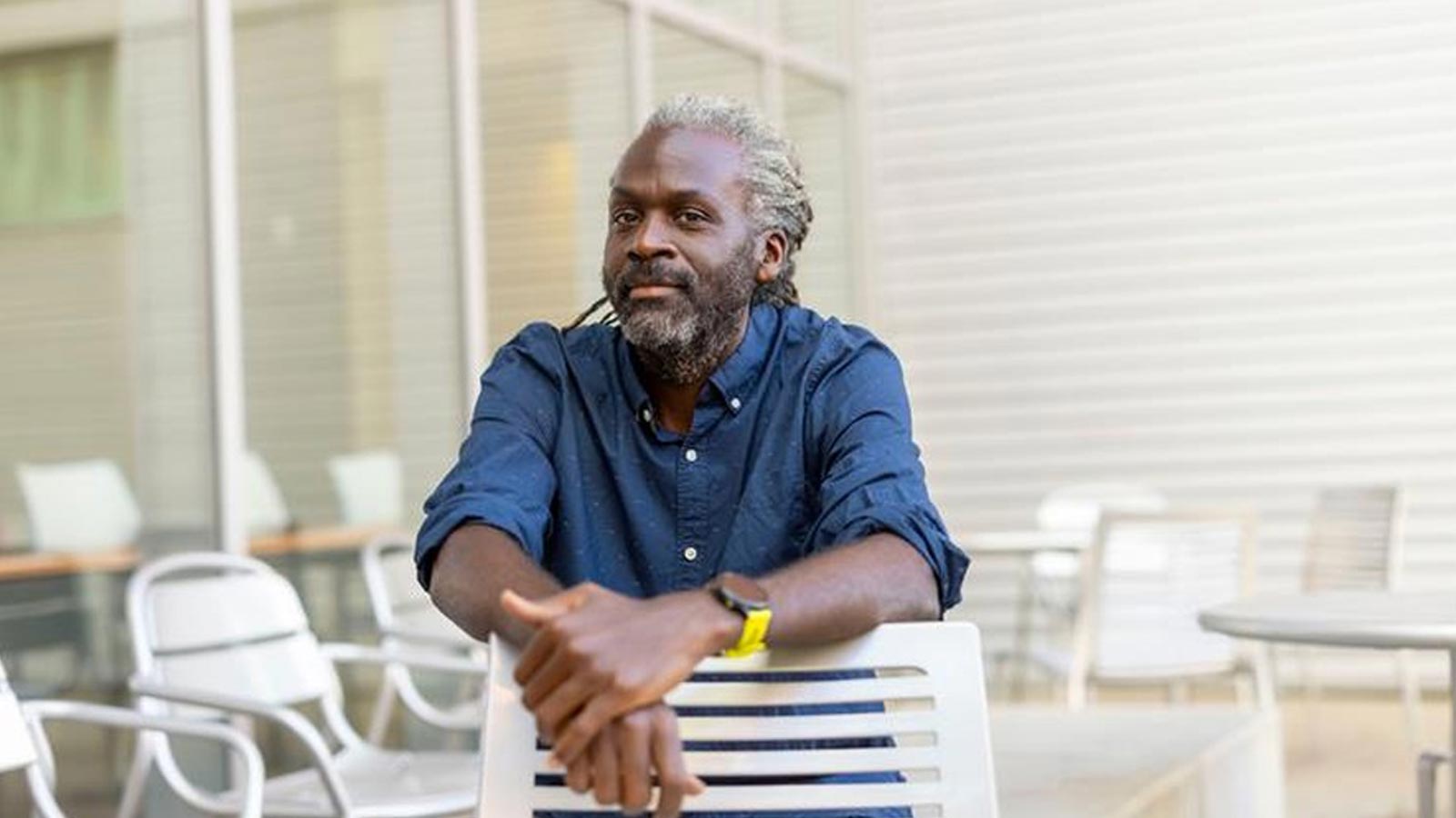 Cal State LA professor Olaseni Sode sits outdoors at a campus patio table, wearing a blue shirt and yellow watch in a relaxed portrait.