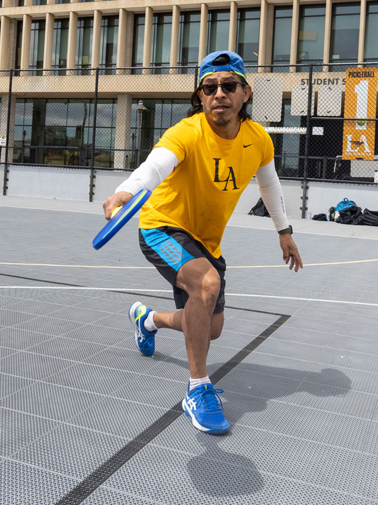 Two people play pickleball on an outdoor court at Cal State LA, with one player in a yellow Cal State LA shirt lunging to hit the ball near the Student Services Building.
